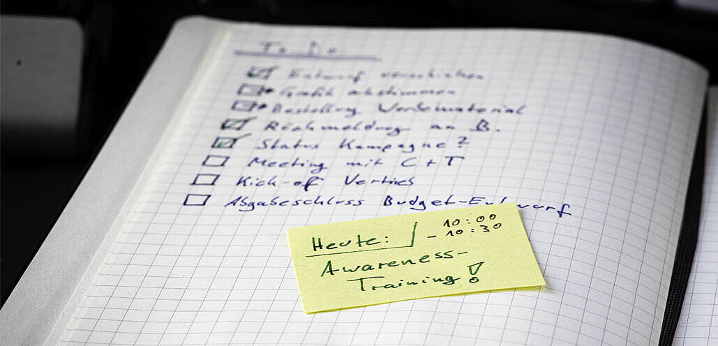 Auf einem karierten Notizbuch steht eine handschriftliche To-do-Liste mit Punkten wie „Kickoff vorbereiten“, „Angebot abstimmen“, „Marketing Maßnahmen“ und „Status Kampagne?“. Darauf klebt ein gelber Zettel mit der Aufschrift „Heute: Awareness-Training!“ sowie der Zeit „10:00 – 10:30“.