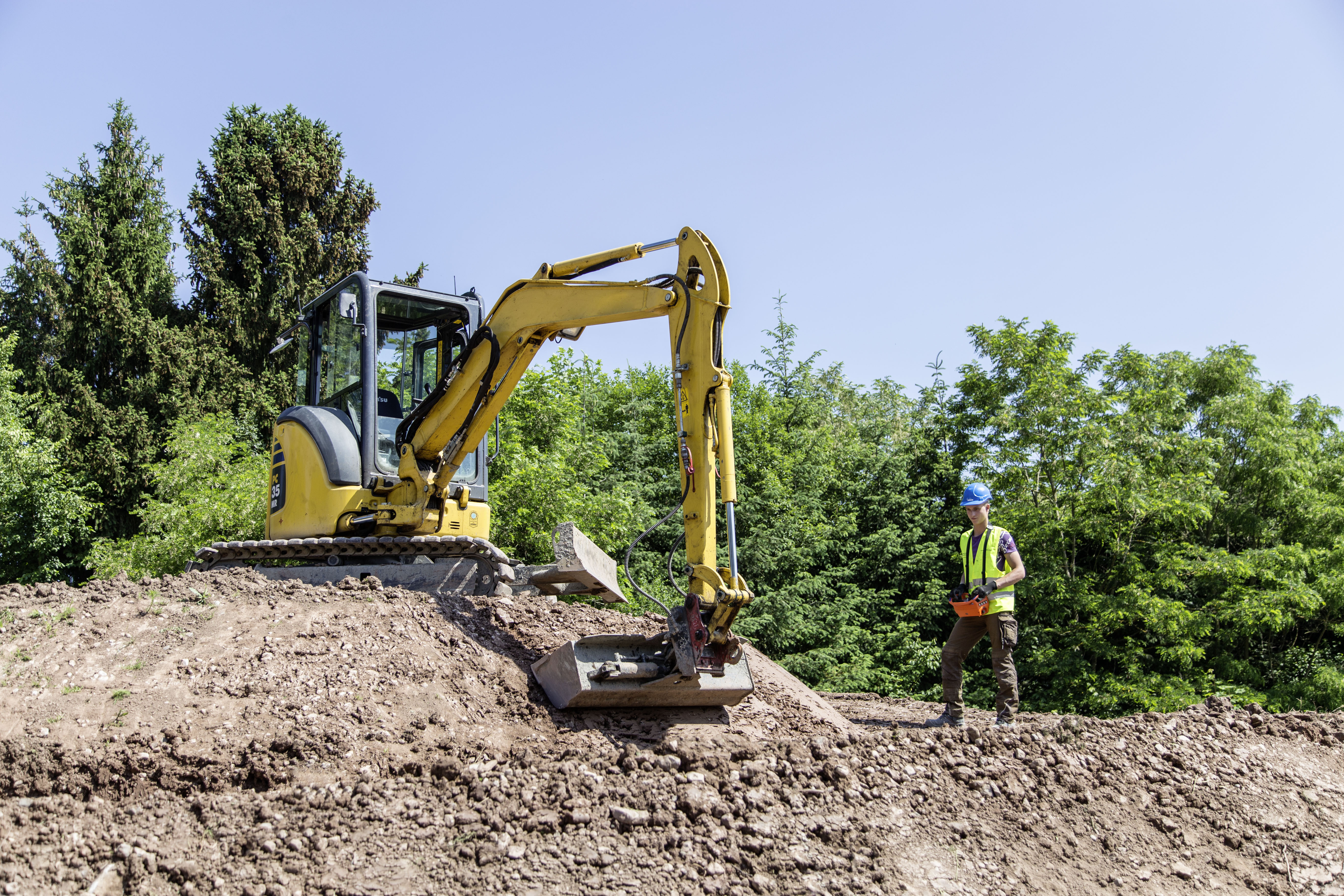 A small yellow tracked excavator stands on a mound of earth, moving its bucket while a construction worker wearing a helmet and high-visibility vest operates it with a remote control. Dense green trees can be seen in the background under a clear blue sky.